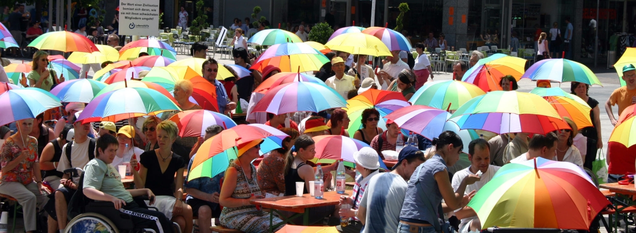 Viele Menschen sitzen in der Sonne an Tischen. Sie halten Schirme in Regenbogenfarben.